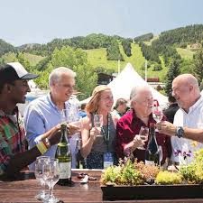 A group of older friends toast with glasses around a table filled with food in a sunny countryside garden setting.