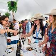 At a conference booth, attendees chat and pose under a tent, including a woman in a straw hat, with badges around their necks.