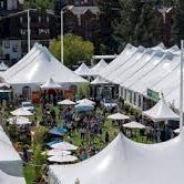 A busy outdoor market with many white canopy tents, stalls, and people in a park or town square.