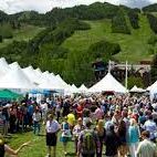 People gather at an outdoor festival with white tents on a sunny field surrounded by green hills.