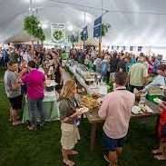 People line up outdoors at a long table to receive food during a community meal, with many standing and chatting.