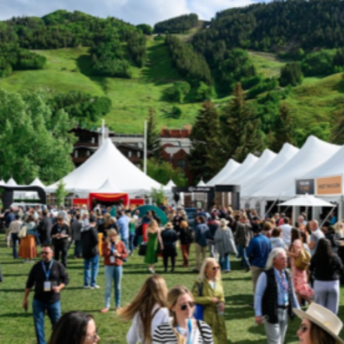 A crowd gathers at an outdoor festival with white tents and booths in a green valley with mountains behind them.