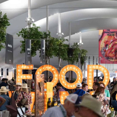 A crowded event under a tent displays large glowing marquee letters reading &ldquo;FOOD&rdquo; with hanging lights and a &ldquo;The Innovators Issue&rdquo; poster.