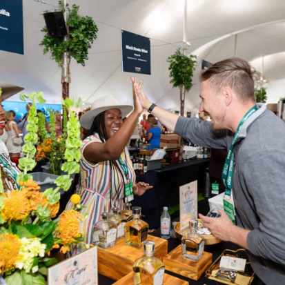 At a trade show, people chat and sample bottled drinks at a decorated booth with lush green-and-orange flowers, smiling and networking.