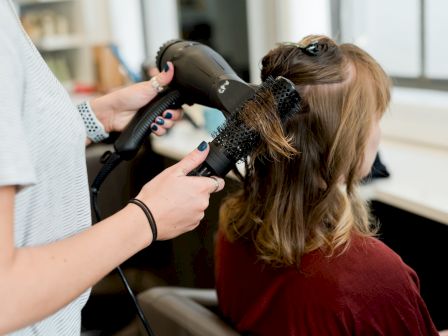 A hairstylist blow-dries a client&rsquo;s long hair with a hair dryer and round brush in a salon.