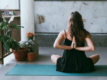 A woman in a black dress sits cross-legged on a yoga mat, hands in prayer, meditating by potted plants and flowers.