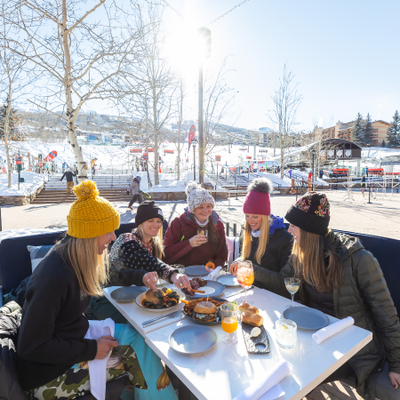 A group of friends in winter clothes enjoy a meal outdoors at a snowy table in a bright park.