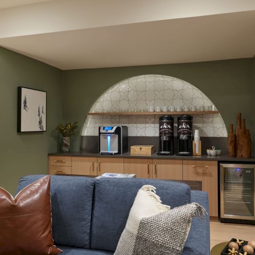 A cozy lounge with a built-in coffee bar&mdash;espresso machines, mugs, and an arched tiled backsplash behind a sofa.