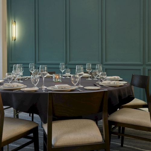 A neatly set dining table with wine glasses, plates, cutlery, and chairs in a modern room with teal paneling.
