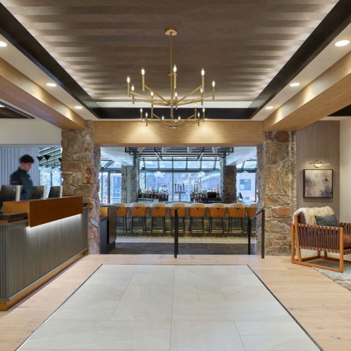 A modern hotel lounge/bar interior with a stone fireplace, brass chandelier, reception desk, and rows of stools behind a counter.