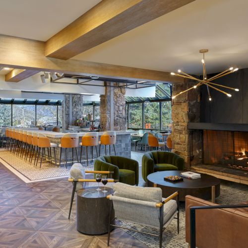 A modern lounge with a stone fireplace, green velvet seating, a round table, and a bar area with stools and large windows.