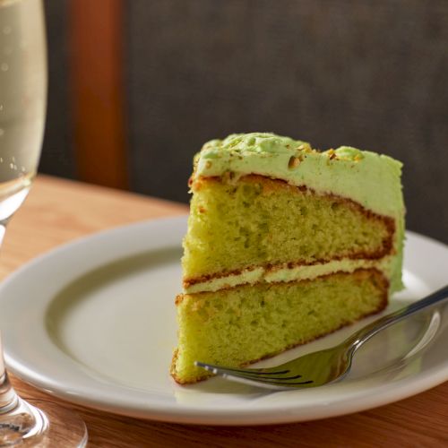 A slice of green pistachio/matcha cake with frosting sits on a plate, with a fork and a champagne flute nearby.