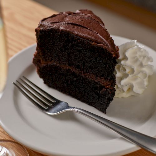 A slice of dark chocolate cake with a layer of frosting, served on a plate with whipped cream and a fork.