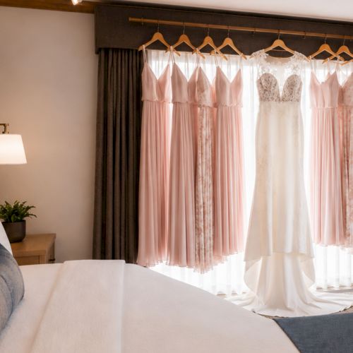A bright hotel bedroom where blush-and-white wedding dresses hang on wooden hangers by the window.