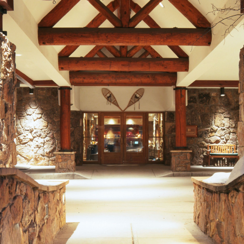 Rustic stone corridor with wooden beams and benches, leading to a lit door under a decorative wing emblem.