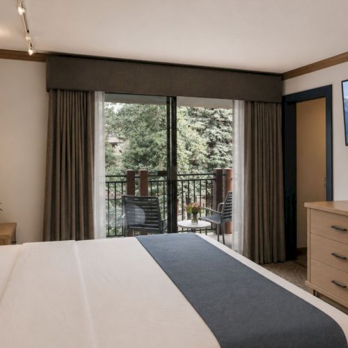 A modern hotel bedroom with a bed, curtains, dresser, and balcony views of mountains through large windows.