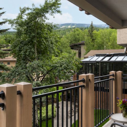 A sunny balcony overlooks lush green trees, distant hills, rooftops, and a glass-covered patio below, for a peaceful outdoor view.