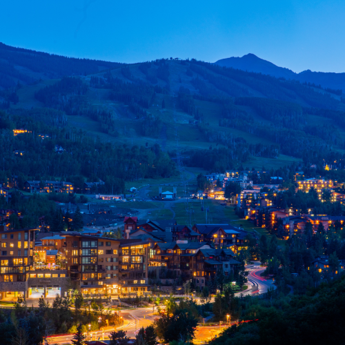 A mountain valley town at dusk, glowing lights along a winding road beneath dark forested hills.
