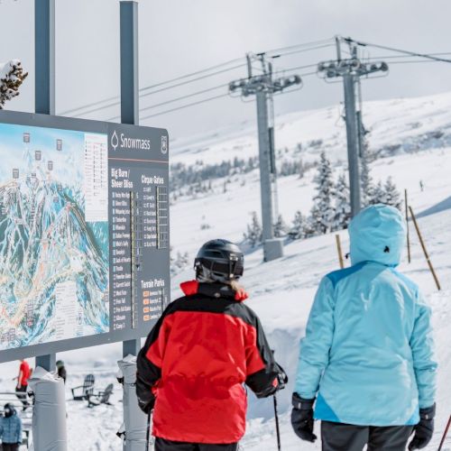Skiers in colorful jackets study a trail map at a snowy ski resort with chairlift towers in the background.