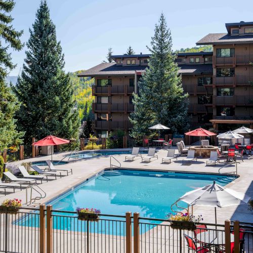 Sunlit apartment complex courtyard with a blue swimming pool, lounge chairs, red umbrellas, and trees.