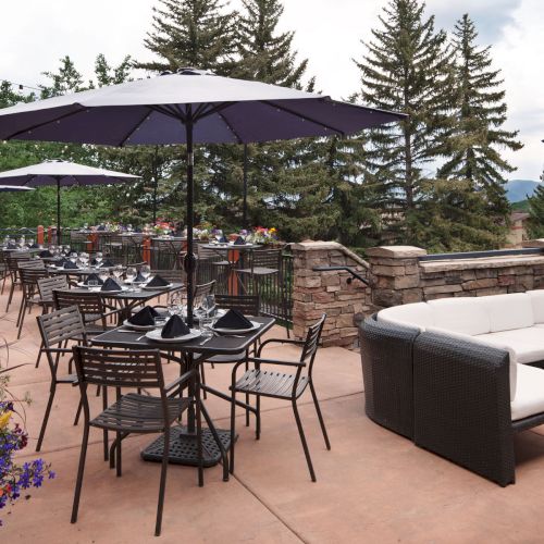 An outdoor patio dining setup with stacked chairs, set tables under dark umbrellas, flowers, and evergreen trees in the background.