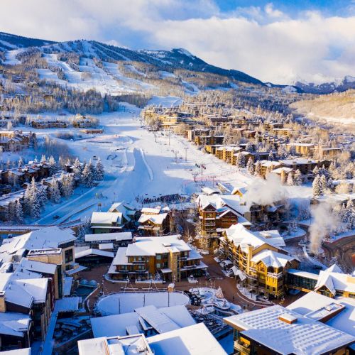 Snowy alpine village with chalets, a ski slope, lift lines, and mountain peaks under a cloudy winter sky.