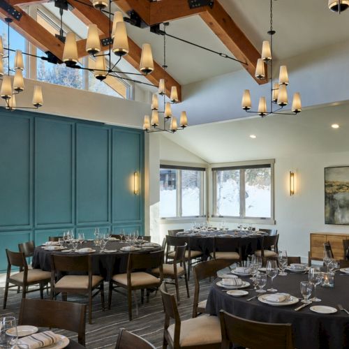 An elegant dining hall with round tables set for guests, warm chandeliers, and teal paneling beneath a wooden vaulted ceiling.