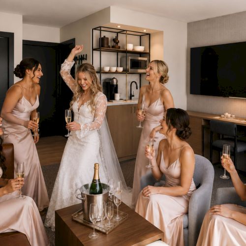 Bridal party in a hotel suite, women in satin dresses toasting champagne and laughing near the kitchenette and TVs.
