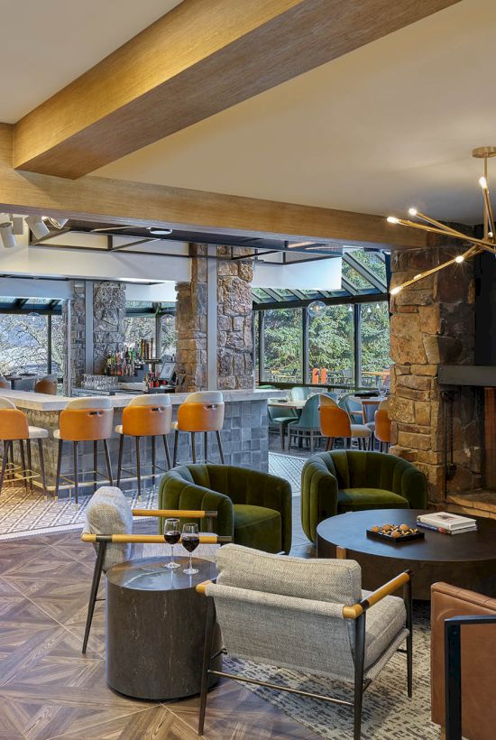 A cozy modern bar lounge with a stone fireplace, green sofas, wooden counter stools, pendant lights, and large windows out to trees.