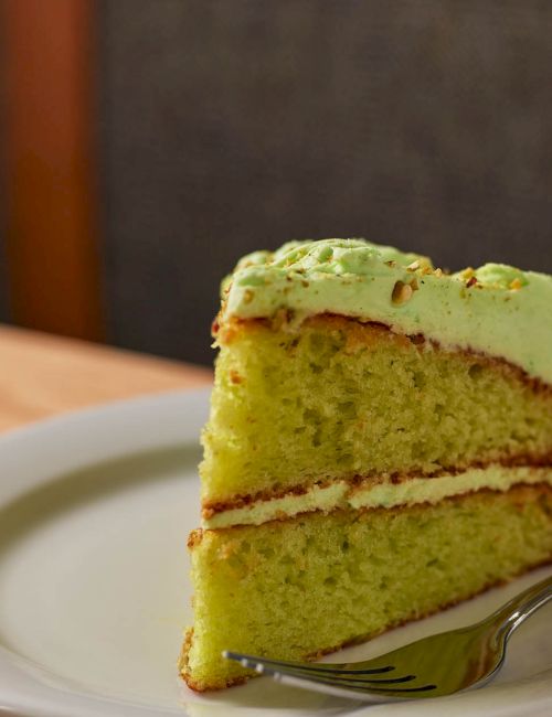 A slice of green tea cake with frosting sits on a plate beside a glass of sparkling drink, ready to enjoy.