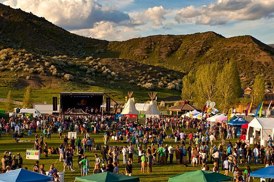 Crowd at an outdoor festival in a valley with tents and a stage, surrounded by green mountains and dramatic clouds.
