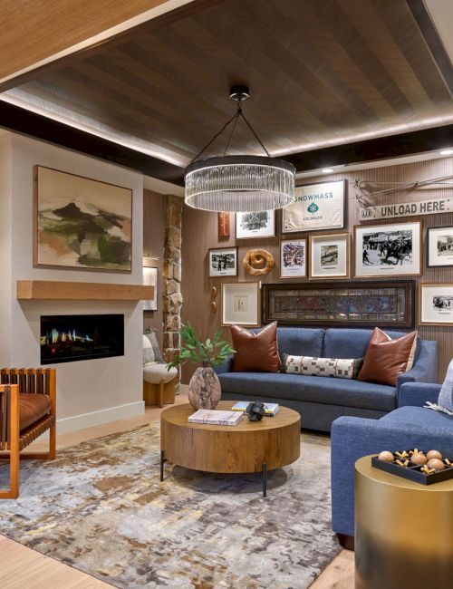 A cozy modern living room with a fireplace, blue sofa, armchairs, framed photos, and a hanging chandelier lighting the space.