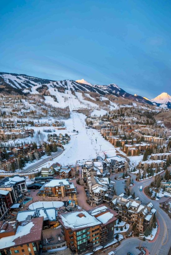 A snowy ski resort village with ski slopes, chairlifts, and houses nestled in a mountain valley under a clear blue sky.