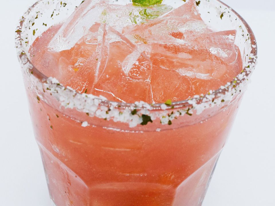 A pink-orange cocktail in a glass, filled with ice, topped with a green lime wedge and sprinkled herb garnish.