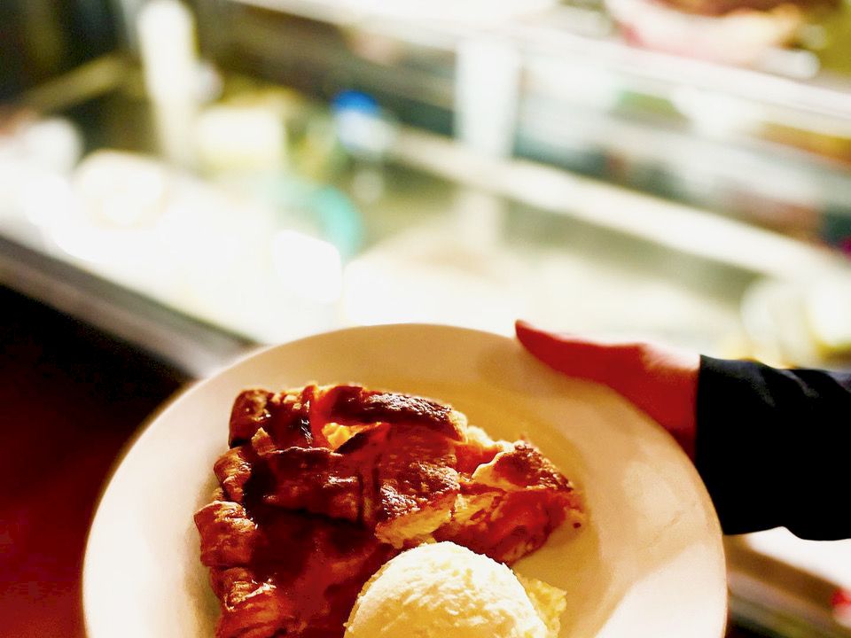 A plate holds a serving of saucy meat with a breaded/roasted potato beside it, outdoors in a sunny blurred setting.