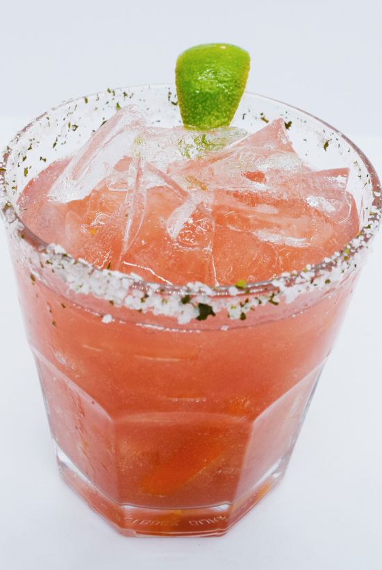 A pink-red cocktail in a glass with ice, a green lime wedge, and a salted/herb rim.