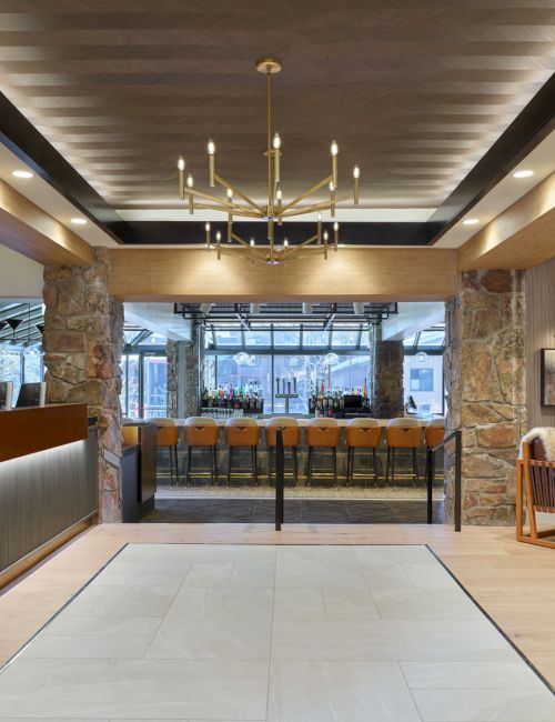 A modern hotel lobby and bar with stone pillars, a chandelier, a long drinks counter, and chairs, plus a receptionist at the desk.